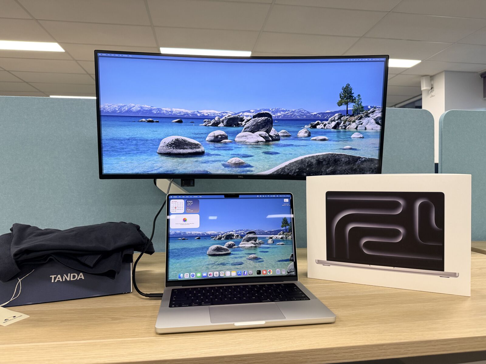 My desk on day one at Tanda: a MacBook Pro hooked up to a curved Dell monitor, the laptop box beside it, and a Tanda box with some merch on top